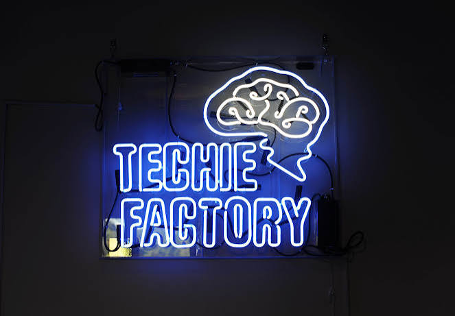 The Techie Factory neon sign glowing blue on a dark wall